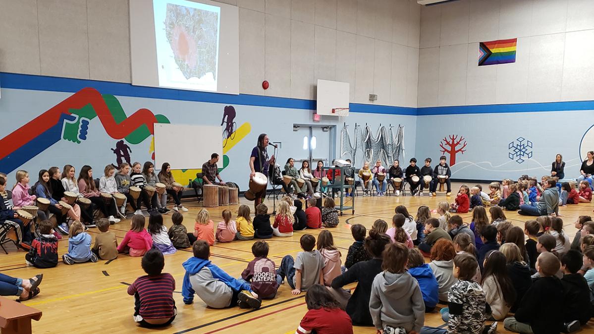 Elementary school students sit cross-legged on a gymnasium floor while a performer plays the Djembe African drum and adults sit in chairs behind him against the gym wall. 