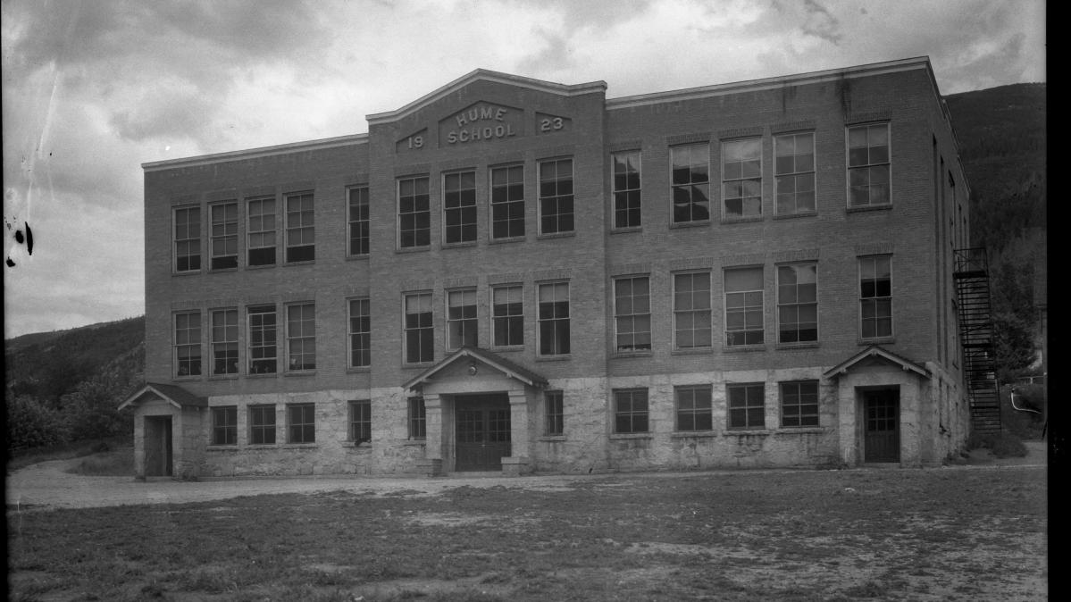 Black and white photograph of the front of a 1930s three story brick schoolhouse with mountains and billowy clouds in the background.