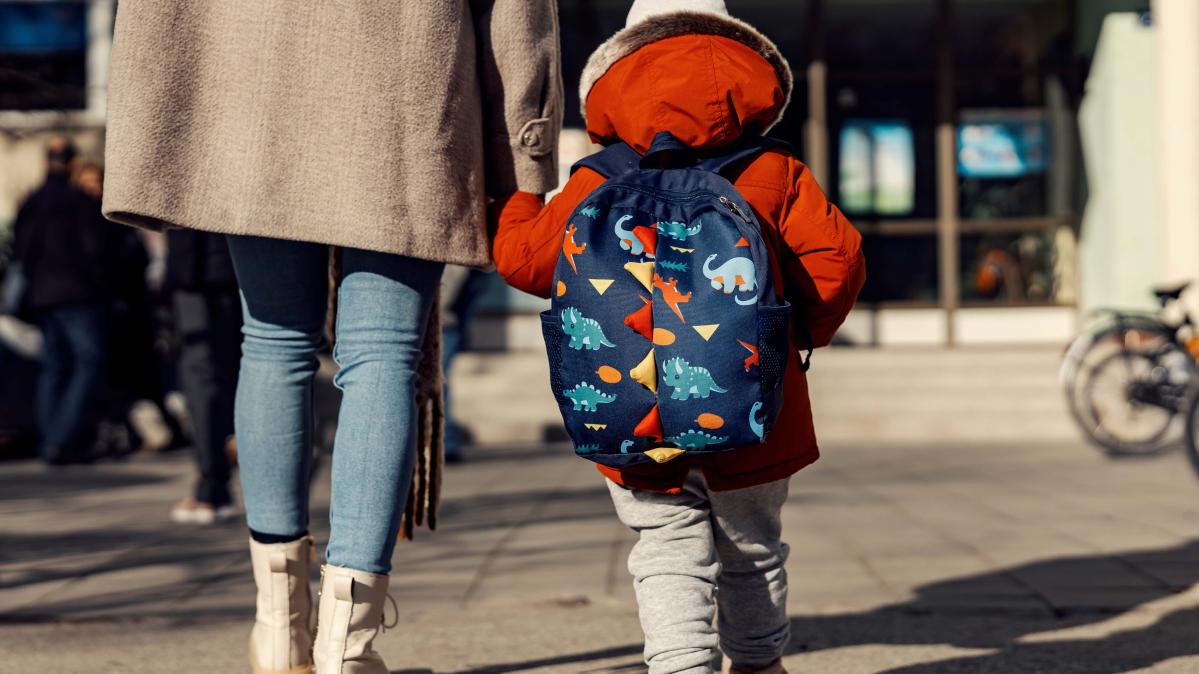 A woman in a beige wool coat walks toward a school with a kindergarten aged child wearing a red jacket and a blue backpack with pictures of animals on it.