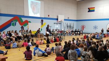 Elementary school students sit cross-legged on a gymnasium floor while a performer plays the Djembe African drum and adults sit in chairs behind him against the gym wall. 