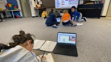 Girl with long brown hair and in a grey sweatshirt lies on a beige carpeted classroom floor writing in a notebook with reference book and open laptop in front of her on the floor. A group of students sit together on the floor across the room in front of a large screen, discussing what's on their laptops and on the screen. 