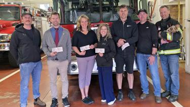 The Nelson Professional Firefighters Charitable Society presented $500 to each of four Nelson area schools to feed students and families in SD8. (l to r) Nelson Professional Firefighter Chas Misener, Wildflower Nelson Principal Nick Graves, Rosemont Elementary Principal Lindsay Mackay, South Nelson Elementary Principal Dawn Snell, Hume Elementary Principal Sacha Kalabis; Nelson Professional Firefighters Chris Thast and Marc Thibault holding firedog Mookie the miniature dachshund. A group of people stand in front of a fire truck inside a fire department, holding cheques in front of themselves.
