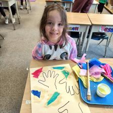 Grade school girl with medium brown hair wearing pink, black and white shirt sits at a school desk in front of a drawing of her hands on beige paper and paint and other art materials beside her.