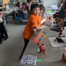 A child in an orange shirt leans over a table in a school classroom while teens stand opposite