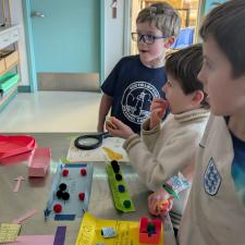 Three boys look at labeled items on a long table in a classroom