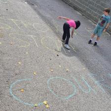 Two children draw words in chalk on a paved access road beside a school