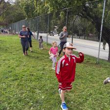 A boy in a ball cap and a red Adidas track top and shorts jogs toward the finish of a school run with parents and other students running across the grass in the background.