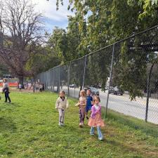 Children jog, saunter and walk through a school field behind a chain link fence with a street in the backgroun