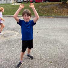 A smiling boy with brown hair wearing a blue t-shirt, black shorts and black running shoes holds his hands above his head while he stands on a paved access road to a school