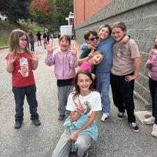 A group of girls and one boy smile and gesture happily in front of a school