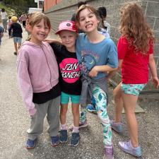 Three girls grimace and smile with their arms around one another while other children walk in the background beside a brick school